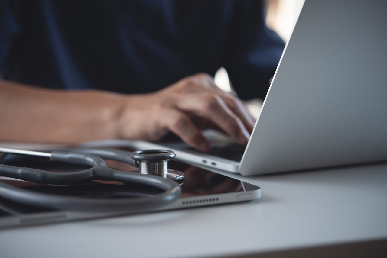 Medizinisches Fachpersonal arbeitet am Laptop mit Stethoskop auf dem Tisch.