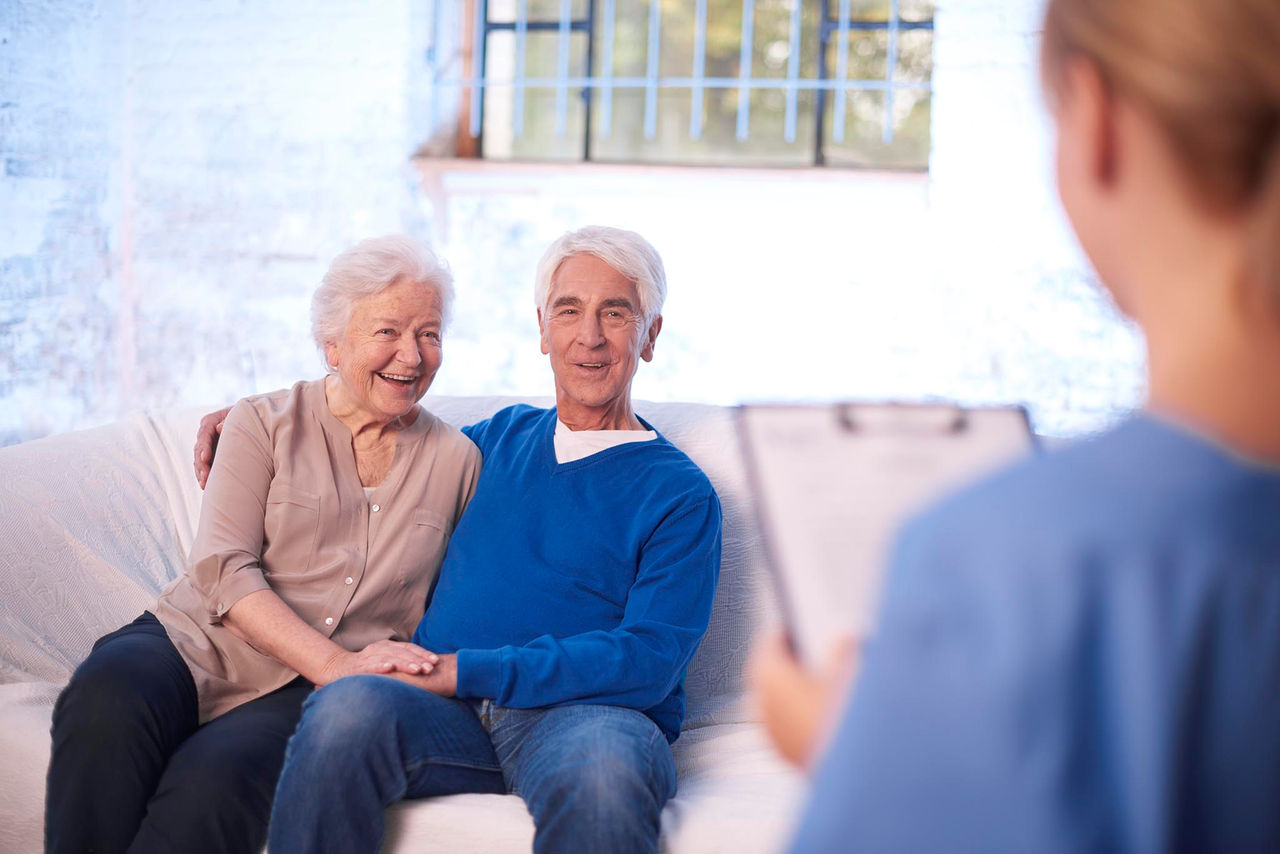 Zwei Personen sitzen nah beieinander auf einem Sofa, während eine betreuende Fachkraft mit Klemmbrett ein Beratungsgespräch in einer medizinischen Umgebung führt.
