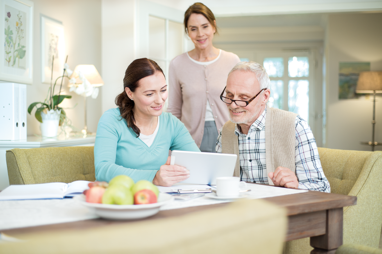 Senior und Pflegefachkraft besprechen Informationen auf einem Tablet, betreut von einer weiteren Fachperson in hellem Wohnambiente.